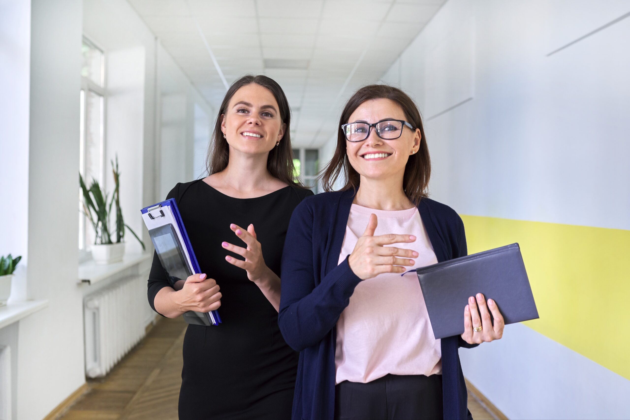 Deux collègues, une femme d’affaires et une enseignante, marchent et discutent dans le couloir d’un bureau ou d’une école. Jeunes et d’âge moyen, elles sourient, illustrant un concept positif de femmes professionnelles dans les domaines des affaires et de l’éducation.